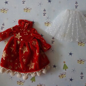 Puede incluir: Un vestido de mu&ntilde;eca de terciopelo rojo con detalles de estrellas doradas y una falda de tul blanco. El vestido tiene mangas largas, cuello alto y est&aacute; adornado con pompones blancos. La falda es blanca con un estampado de lunares.