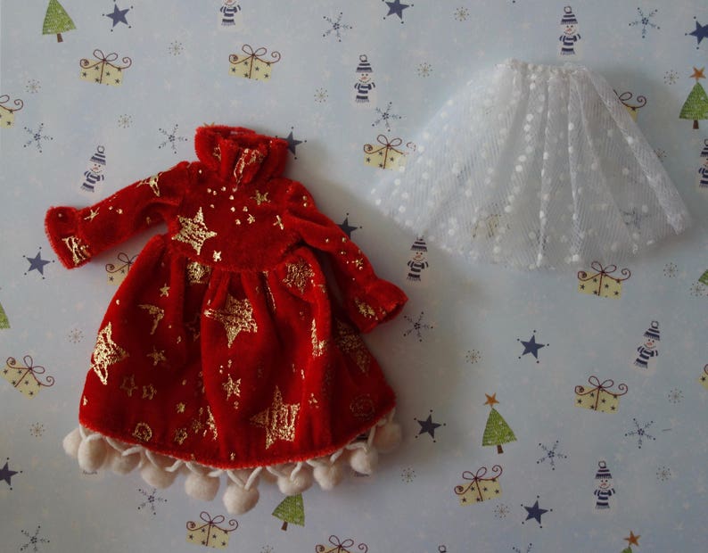 Puede incluir: Un vestido de mu&ntilde;eca de terciopelo rojo con detalles de estrellas doradas y una falda de tul blanco. El vestido tiene mangas largas, cuello alto y est&aacute; adornado con pompones blancos. La falda es blanca con un estampado de lunares.