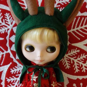 Puede incluir: Una mu&ntilde;eca vestida con un vestido rojo festivo con detalles de estrellas doradas y una capucha verde con cuernos de fieltro marr&oacute;n. La mu&ntilde;eca est&aacute; sobre un fondo rojo con un patr&oacute;n de copo de nieve blanco, creando un tema navide&ntilde;o.