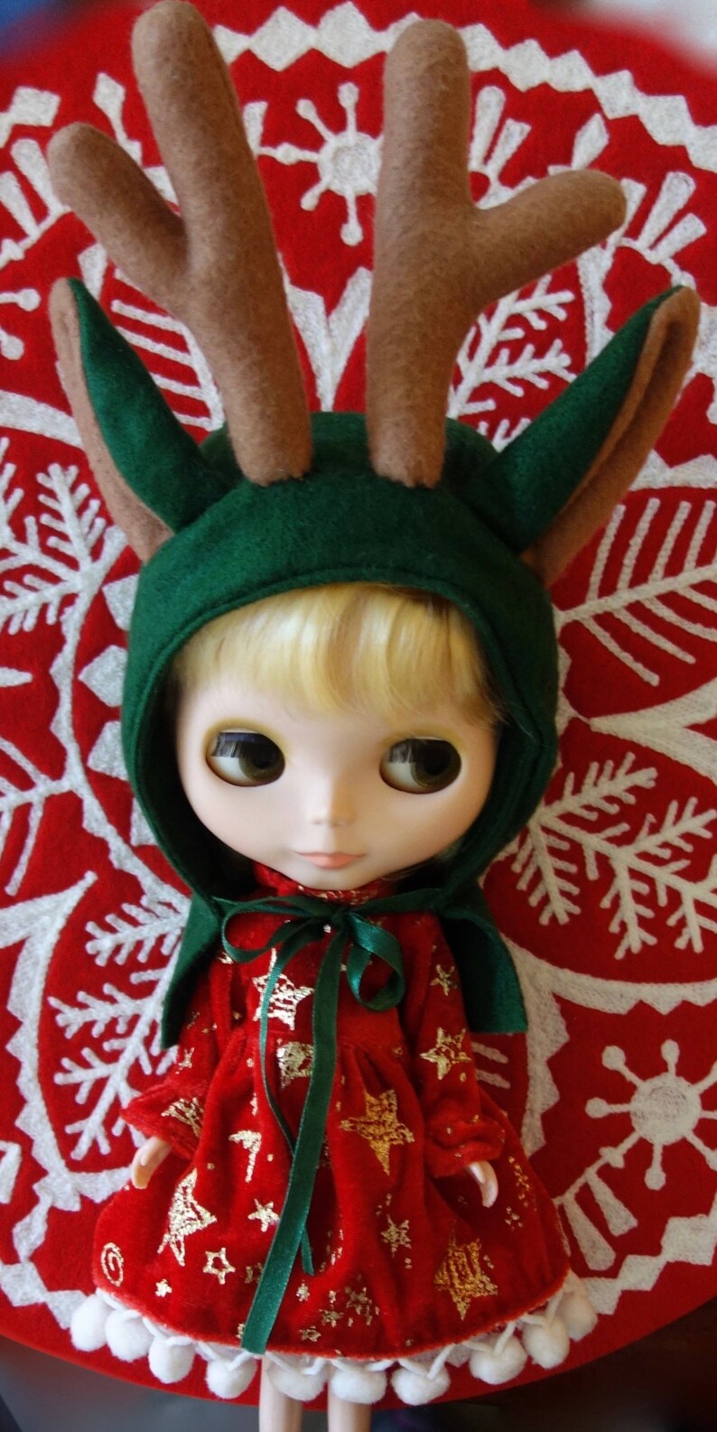 Puede incluir: Una mu&ntilde;eca vestida con un vestido rojo festivo con detalles de estrellas doradas y una capucha verde con cuernos de fieltro marr&oacute;n. La mu&ntilde;eca est&aacute; sobre un fondo rojo con un patr&oacute;n de copo de nieve blanco, creando un tema navide&ntilde;o.