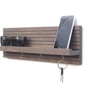May include: A brown wooden wall mounted key holder with a shelf, hooks, and a phone slot. Sunglasses are on the shelf, and keys are on a hook.