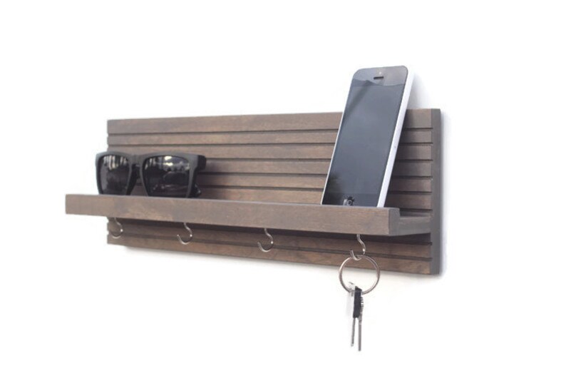 May include: A brown wooden wall mounted key holder with a shelf, hooks, and a phone slot. Sunglasses are on the shelf, and keys are on a hook.