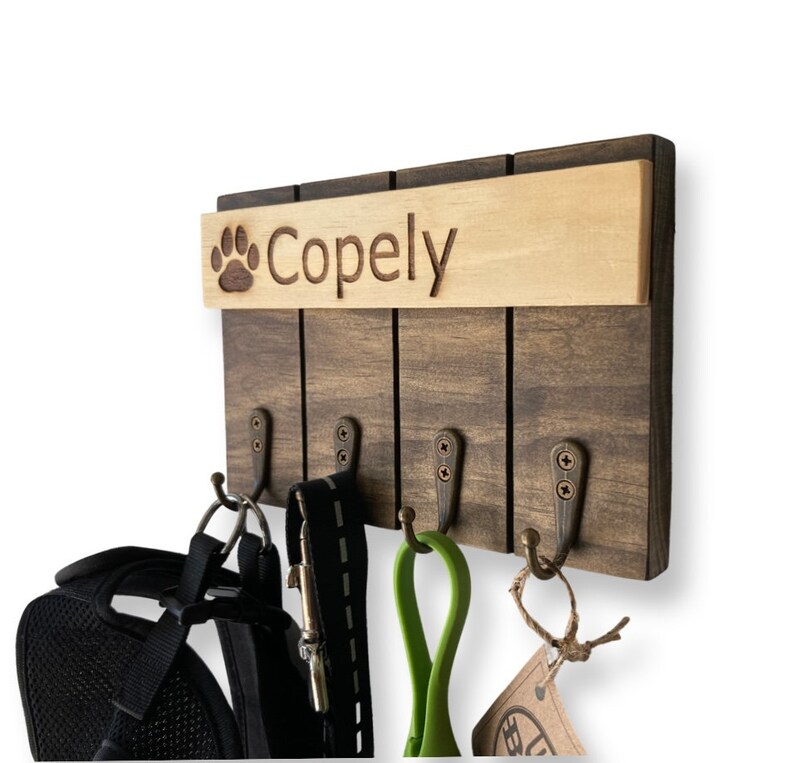 May include: A wooden dog leash and collar rack with four hooks. The rack has a brown stain and a light wood top with the name "Copely" and a paw print engraved on it.