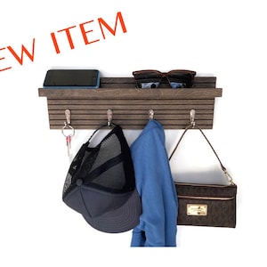 May include: A wooden wall mounted key holder with a shelf on top. The key holder has four hooks and a slatted design. There is a cell phone, sunglasses, a baseball cap, a blue jacket, and a brown purse hanging on the key holder.