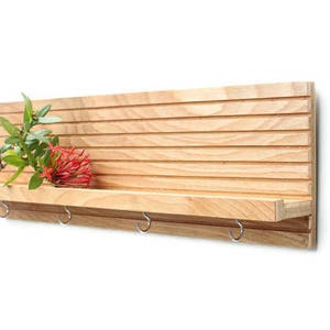 Pu&ograve; includere: Un ripiano a muro in legno con un design a doghe e un ripiano con quattro ganci. Il ripiano &egrave; in legno chiaro e ha una finitura naturale. Un fiore rosso &egrave; sul ripiano.