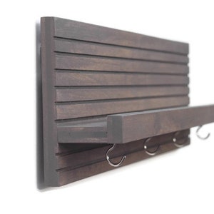 May include: A dark brown wooden wall shelf with a small shelf and three hooks. The shelf is mounted on a white wall.