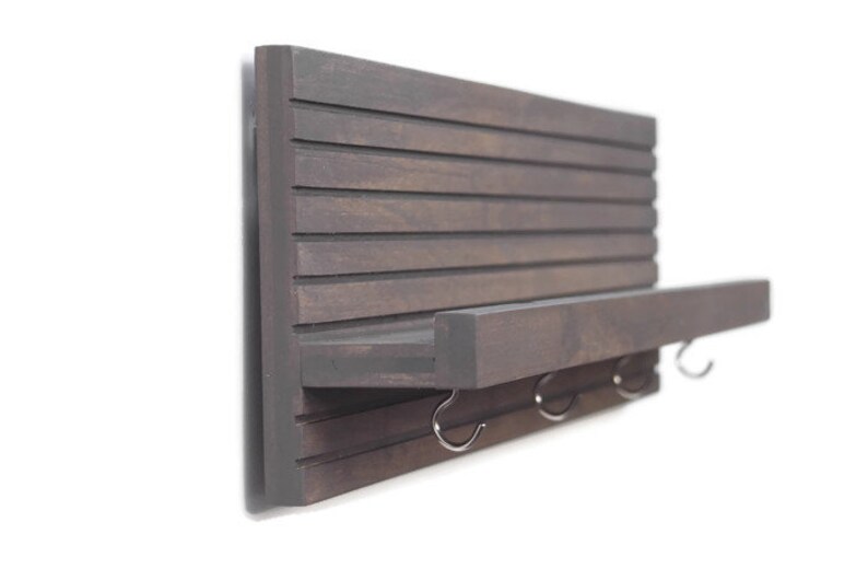 May include: A dark brown wooden wall shelf with a small shelf and three hooks. The shelf is mounted on a white wall.