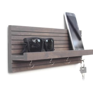May include: A wooden wall-mounted key holder with a shelf, hooks, and a phone slot. The key holder is painted a dark brown color and has a slatted design. Sunglasses are on the shelf, and a key is on one of the hooks.