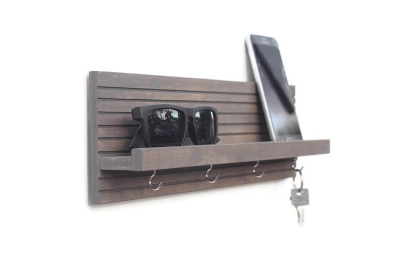 May include: A wooden wall-mounted key holder with a shelf, hooks, and a phone slot. The key holder is painted a dark brown color and has a slatted design. Sunglasses are on the shelf, and a key is on one of the hooks.
