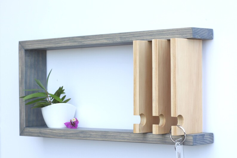 Puede incluir: Una estanter&iacute;a de pared de madera gris con una peque&ntilde;a planta en maceta y una flor en la estanter&iacute;a. La estanter&iacute;a tiene un porta llaves con cuatro ranuras y una llave colgando.