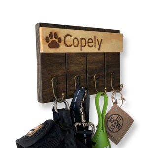 May include: Wooden dog leash and key holder with four hooks. The holder has a brown wood finish and a top panel with a paw print and the name "Copely" engraved in it.