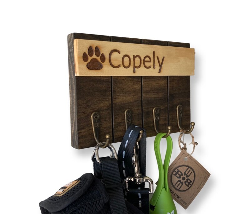 May include: Wooden dog leash and key holder with four hooks. The holder has a brown wood finish and a top panel with a paw print and the name "Copely" engraved in it.