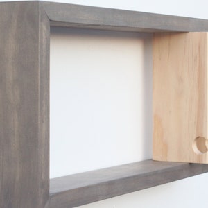 May include: A gray wooden wall shelf with a light wood shelf insert that has three cut-out hooks. The shelf is mounted on a white wall.