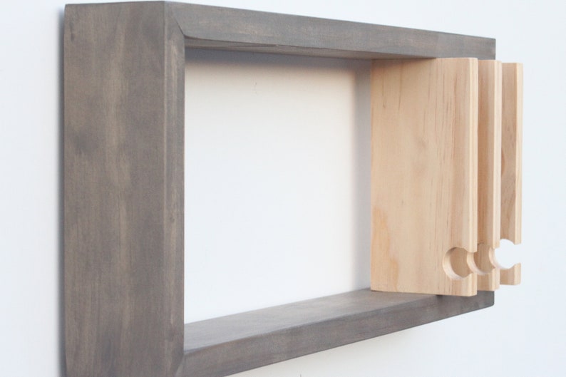 May include: A gray wooden wall shelf with a light wood shelf insert that has three cut-out hooks. The shelf is mounted on a white wall.