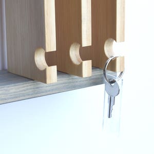 Puede incluir: Un porta llaves de madera con cuatro ranuras para llaves. El porta llaves est&aacute; montado en una repisa de madera gris. Una sola llave cuelga del porta llaves.