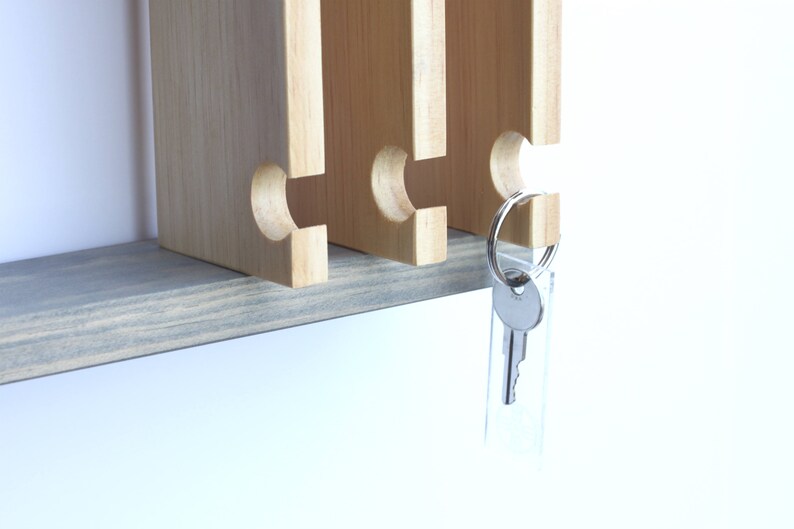 Puede incluir: Un porta llaves de madera con cuatro ranuras para llaves. El porta llaves est&aacute; montado en una repisa de madera gris. Una sola llave cuelga del porta llaves.