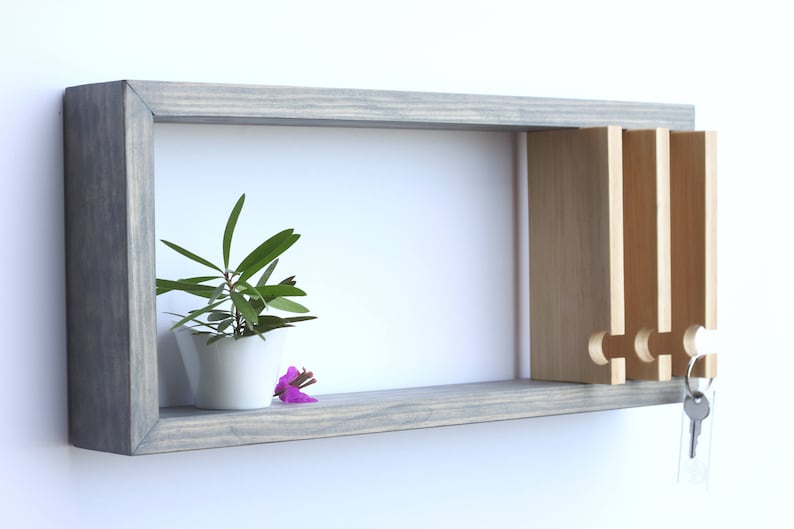 Puede incluir: Una estanter&iacute;a de pared de madera gris con una peque&ntilde;a planta en maceta y un colgador de llaves con cuatro ganchos. El colgador de llaves est&aacute; hecho de madera clara y tiene un dise&ntilde;o decorativo.
