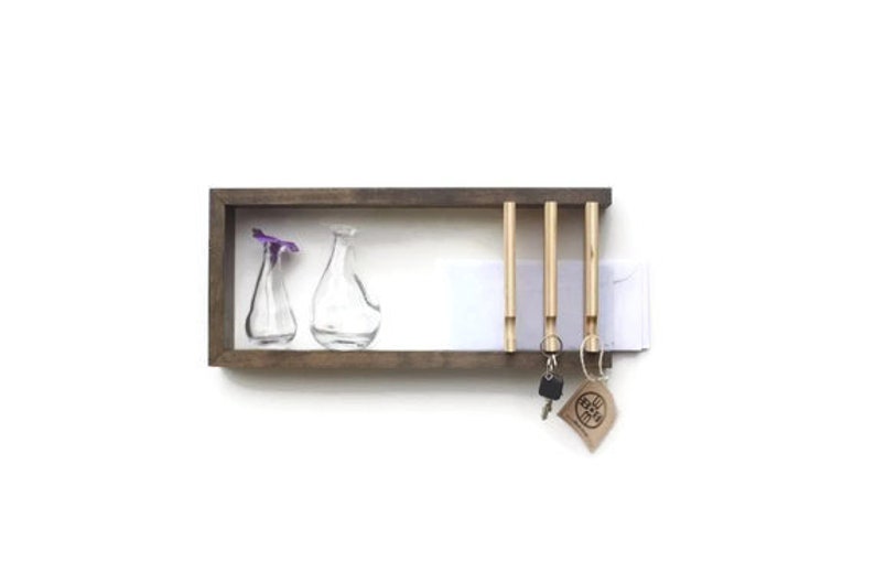 May include: A wooden key holder with four hooks and a small shelf. The shelf has two clear glass vases with purple flowers. The key holder is mounted on a white wall.