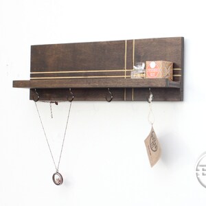 May include: A dark brown wooden wall shelf with gold accents and three hooks. A necklace hangs from one hook, and a small box sits on the shelf. The shelf is mounted on a white wall.
