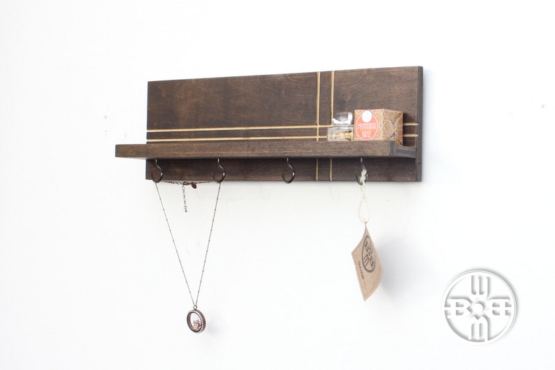 May include: A dark brown wooden wall shelf with gold accents and three hooks. A necklace hangs from one hook, and a small box sits on the shelf. The shelf is mounted on a white wall.