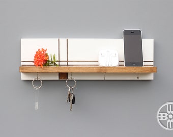 May include: White wall mounted key holder with a shelf and hooks. The shelf has a white background with brown lines and a wooden front. There are two sets of keys hanging from the hooks. A cell phone and a white case with earbuds are on the shelf. A small orange flower is on the shelf.