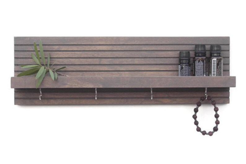 May include: A gray wooden shelf with a slatted back and a small shelf on the front. There are five small bottles on the top shelf and a beaded necklace hanging from a hook on the bottom shelf.