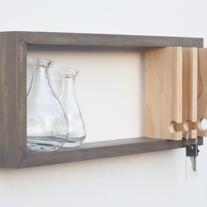 May include: A gray wooden wall shelf with a key holder on the right side. The shelf has a small open space for displaying items, and two clear glass vases are sitting on the shelf.