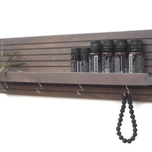 May include: A dark brown wooden wall shelf with a small shelf and hooks. The shelf has a slatted design and is holding several small bottles. A black beaded necklace hangs from one of the hooks.