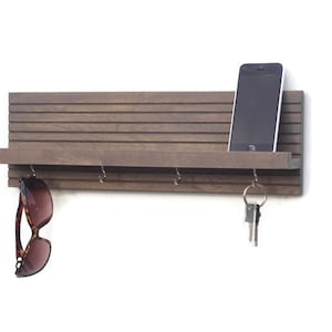 May include: A gray wooden wall mounted key holder with a shelf and hooks. A cell phone is on the shelf and sunglasses are hanging from a hook.