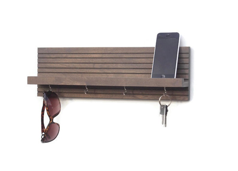 May include: A gray wooden wall mounted key holder with a shelf and hooks. A cell phone is on the shelf and sunglasses are hanging from a hook.