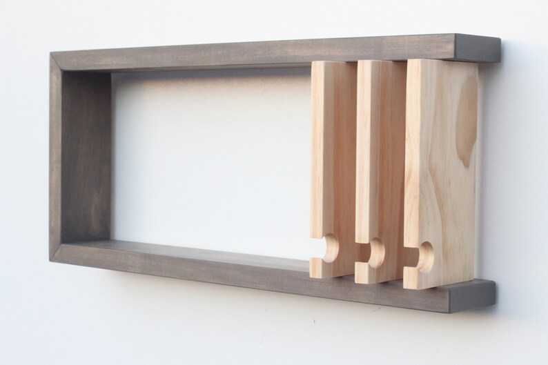 May include: A gray wooden wall shelf with a rectangular opening and five wooden pegs protruding from the side. The pegs have a rounded, curved shape at the bottom.