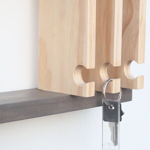 May include: A wooden key holder with three slots for keys. The key holder is mounted on a dark gray shelf. A key with a silver key ring is hanging from the holder.