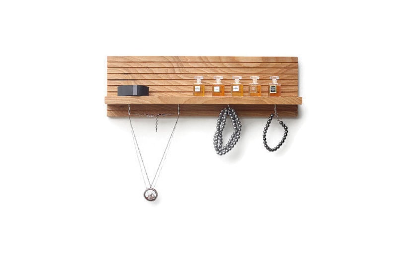 Pu&ograve; includere: Un ripiano a muro in legno con un design a doghe. Il ripiano ha una scatola nera sullo scaffale superiore e cinque piccole bottiglie di vetro sullo scaffale intermedio. Ci sono due collane e un braccialetto appesi a ganci sotto il ripiano.