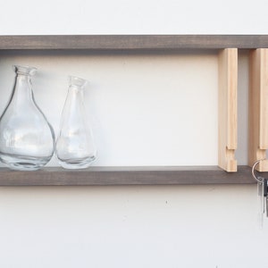 May include: A gray wooden wall shelf with a key holder and two glass vases. The key holder has four slots for keys and is made of light wood. The shelf has a simple, modern design.