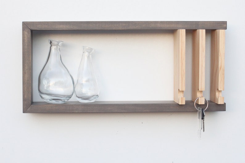 May include: A gray wooden wall shelf with a key holder and two glass vases. The key holder has four slots for keys and is made of light wood. The shelf has a simple, modern design.