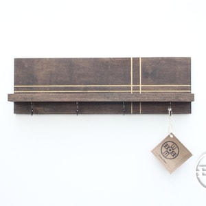 May include: A dark brown wooden wall shelf with a gold stripe design. The shelf has four hooks and a small shelf on top. A brown paper tag with the word "Bohemian" is hanging from one of the hooks.