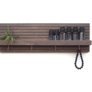 May include: A dark brown wooden shelf with a small plant on the left side. The shelf has a small ledge and hooks on the bottom. There are eight small bottles on the shelf and a black beaded necklace hanging from one of the hooks.