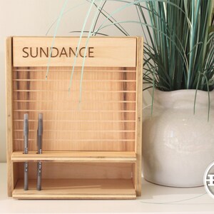 May include: A wooden organizer with the word "SUNDANCE" engraved on the top. The organizer has a shelf and slots for holding pens or pencils. The organizer is sitting on a white surface.