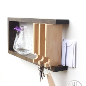 May include: A wooden wall-mounted key holder with a shelf and a small vase. The key holder has a slot for mail and hooks for keys. The vase is clear glass and holds purple flowers.