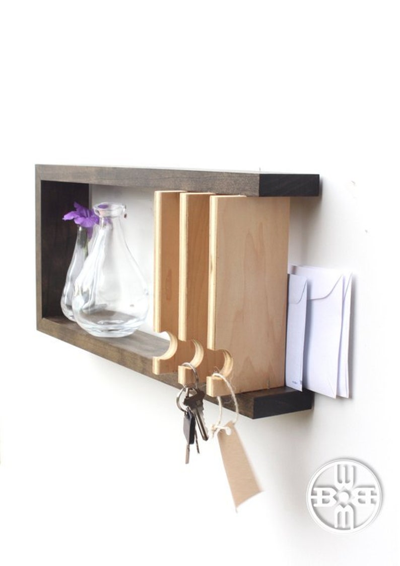May include: A wooden wall-mounted key holder with a shelf and a small vase. The key holder has a slot for mail and hooks for keys. The vase is clear glass and holds purple flowers.