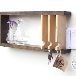 May include: A wooden wall-mounted key holder with a shelf and three key hooks. The shelf has two clear glass vases with purple flowers. The key holder is stained a dark brown color. The key hooks are made of light wood and have a rounded shape. There is a small white tag hanging from the bottom of the key holder with the text "BOOM" on it.