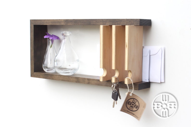 May include: A wooden wall-mounted key holder with a shelf and three key hooks. The shelf has two clear glass vases with purple flowers. The key holder is stained a dark brown color. The key hooks are made of light wood and have a rounded shape. There is a small white tag hanging from the bottom of the key holder with the text "BOOM" on it.