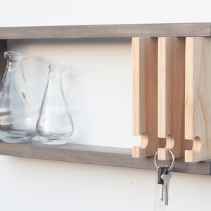 May include: A gray wooden wall shelf with a key holder and two glass vases. The key holder has four slots for keys and is made of natural wood. The shelf has a simple, modern design.