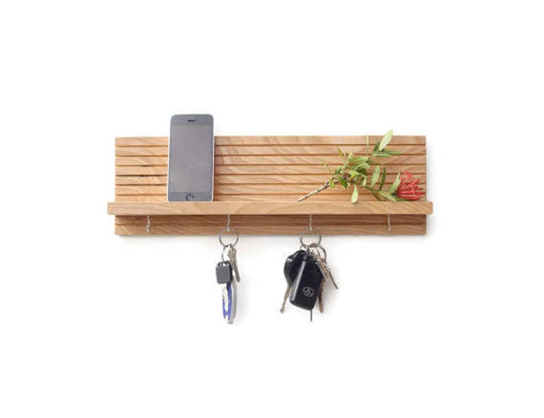 Pu&ograve; includere: Un porta chiavi da parete in legno con un ripiano per un telefono o altri piccoli oggetti. Il porta chiavi ha cinque ganci ed &egrave; realizzato in legno marrone chiaro. C'&egrave; una piccola pianta con fiori rossi sullo scaffale.