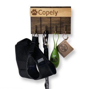 May include: A wooden dog leash and collar rack with four hooks. The rack is personalized with the name "Copely" and a paw print. A black dog harness, a green poop bag dispenser, and a brown tag are hanging from the hooks.