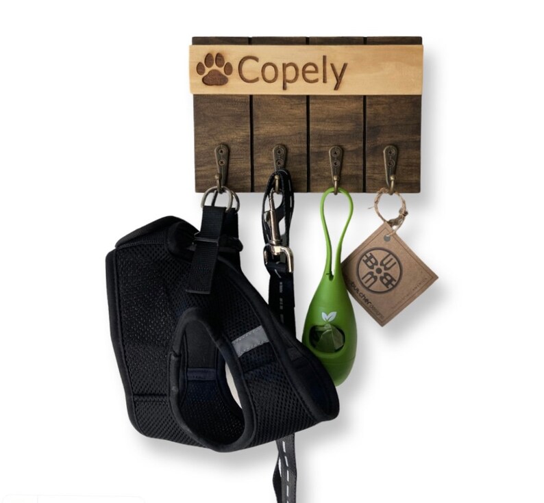 May include: A wooden dog leash and collar rack with four hooks. The rack is personalized with the name "Copely" and a paw print. A black dog harness, a green poop bag dispenser, and a brown tag are hanging from the hooks.