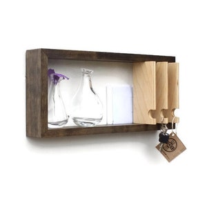 May include: A wooden key holder with a shelf for displaying small items. The key holder has a dark brown frame and a light wood interior. There are three key hooks and two glass vases on the shelf. The key holder is mounted on a white wall.
