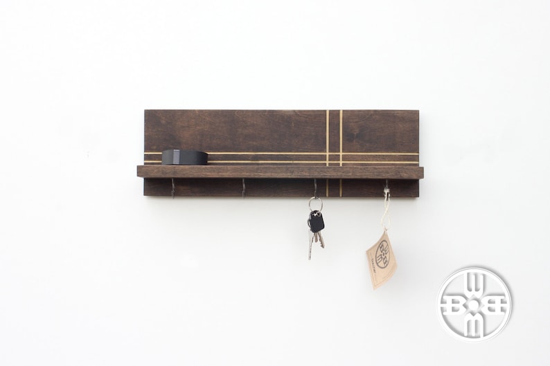 May include: A dark brown wooden key holder with a shelf and gold accents. The key holder has four hooks and a small shelf on top. There are keys and a tag hanging from the hooks.