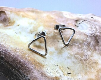 May include: A pair of silver-colored triangle stud earrings. The earrings are displayed on a light brown, textured surface. The earrings have a simple, geometric design, with a triangular shape. The posts are visible on the back of the earrings.