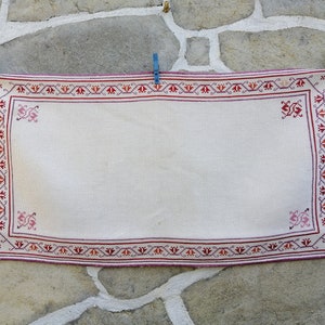 Può includere: Un tappeto bianco tessuto a mano con un bordo floreale rosso e rosa. Il tappeto è appeso a un muro di pietra.